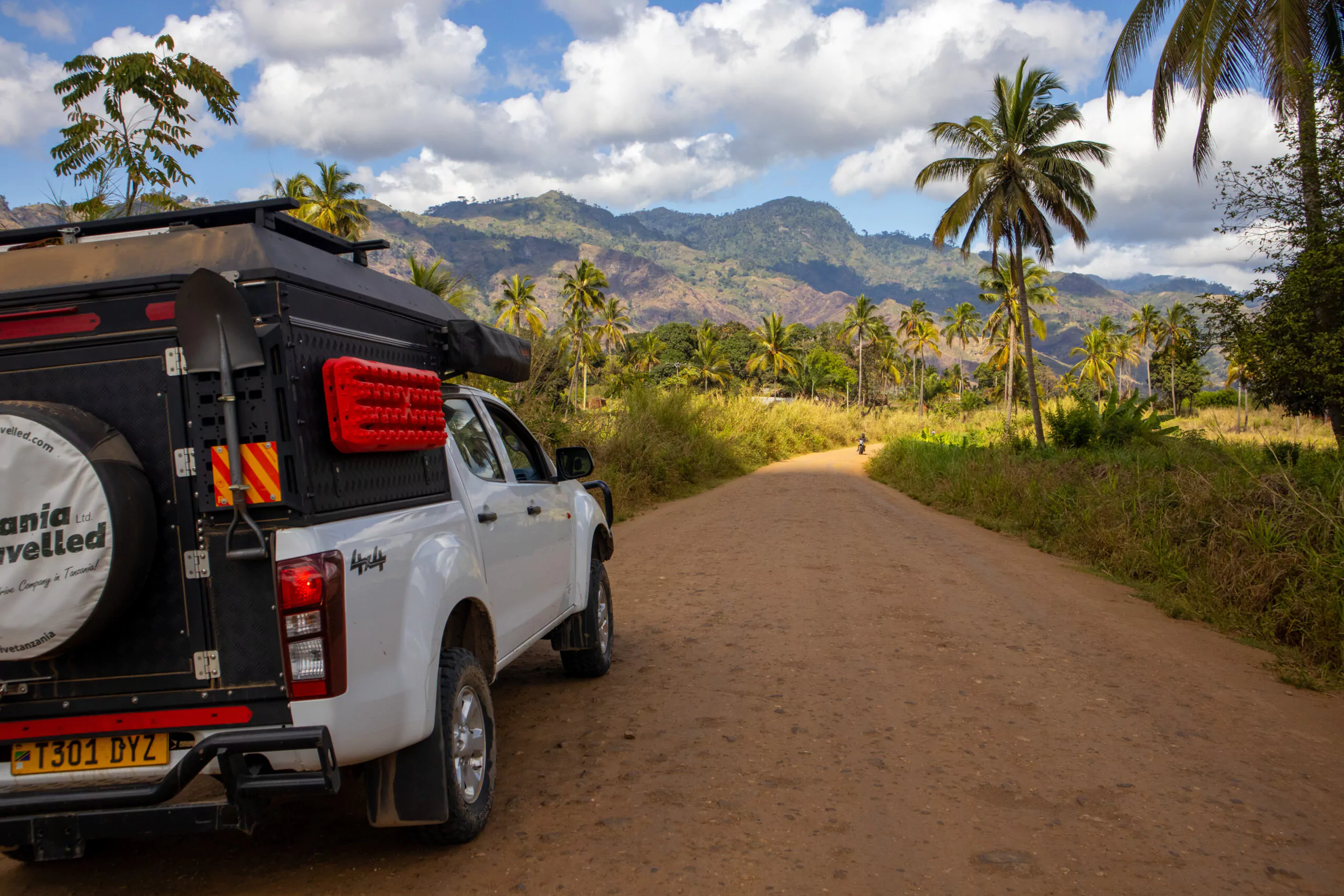 Tansania Camping Safari mit dem Mietwagen Fahrzeug auf Strasse mit Palmen und Berglandschaft